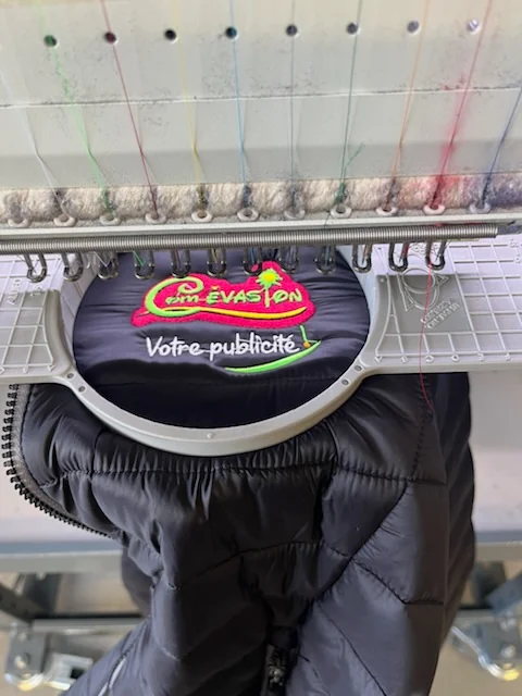 Machine à broder de l’atelier brodant le logo Com Évasion « Votre publicité ! » sur un bodywarmer noir