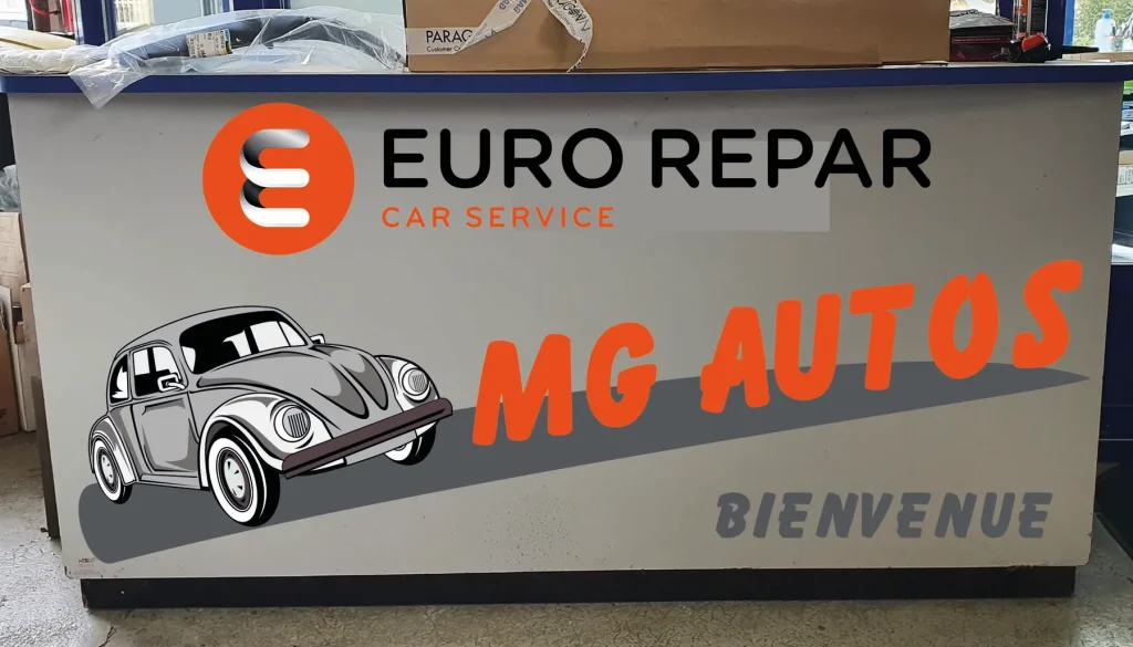 Banque d’accueil habillée d’un panneau gris : logo Eurorepar Car Service, Coccinelle et « MG Autos, Bienvenue »