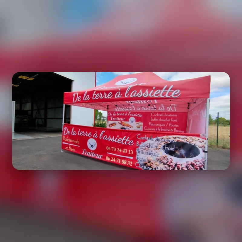 Barnum rouge « De la terre à l’assiette, traiteur » monté devant un hangar
