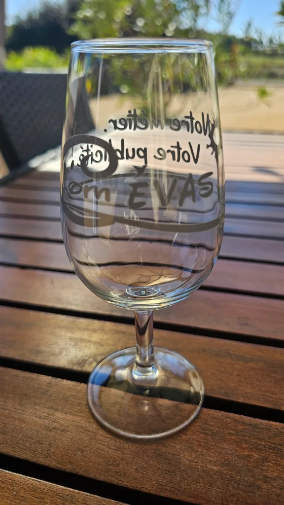 Verre à vin marqué « Votre métier, votre publicité, Com Évasion » posé sur une table en bois