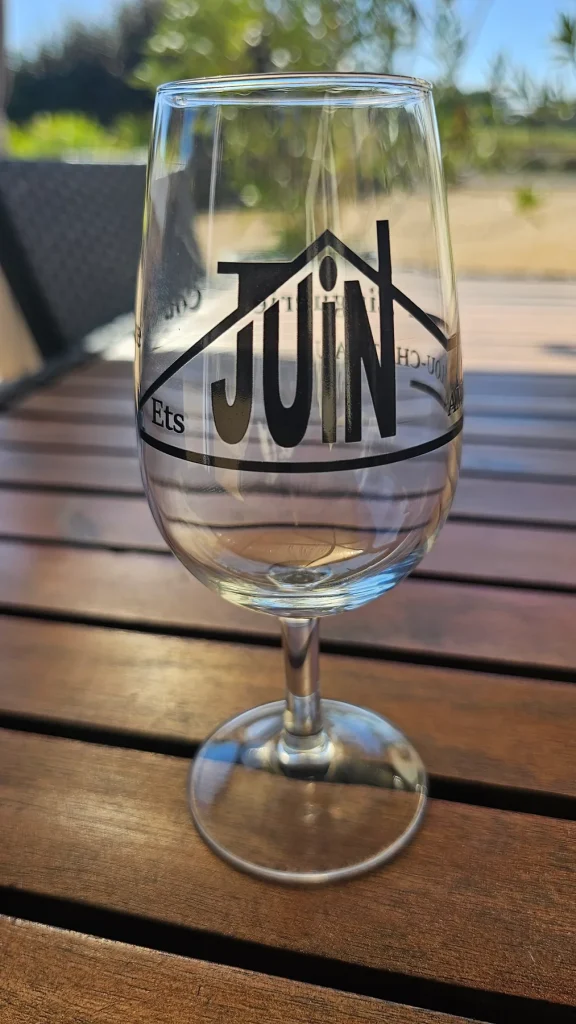 Verre à vin marqué en noir du logo Ets Juin, posé sur une table en bois au soleil