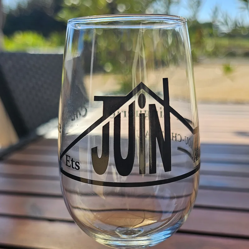 Verre à vin marqué en noir du logo Ets Juin, posé sur une table en bois au soleil