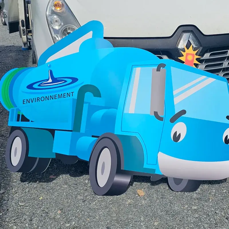 Panneau Dibond découpé en forme de camion de vidange bleu souriant, posé devant un utilitaire