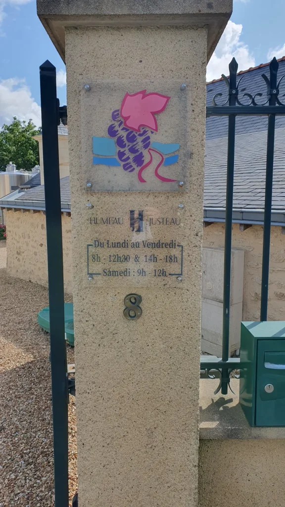 Plaque transparente avec une grappe de raisin et les horaires du domaine Humeau Justeau sur un pilier en pierre